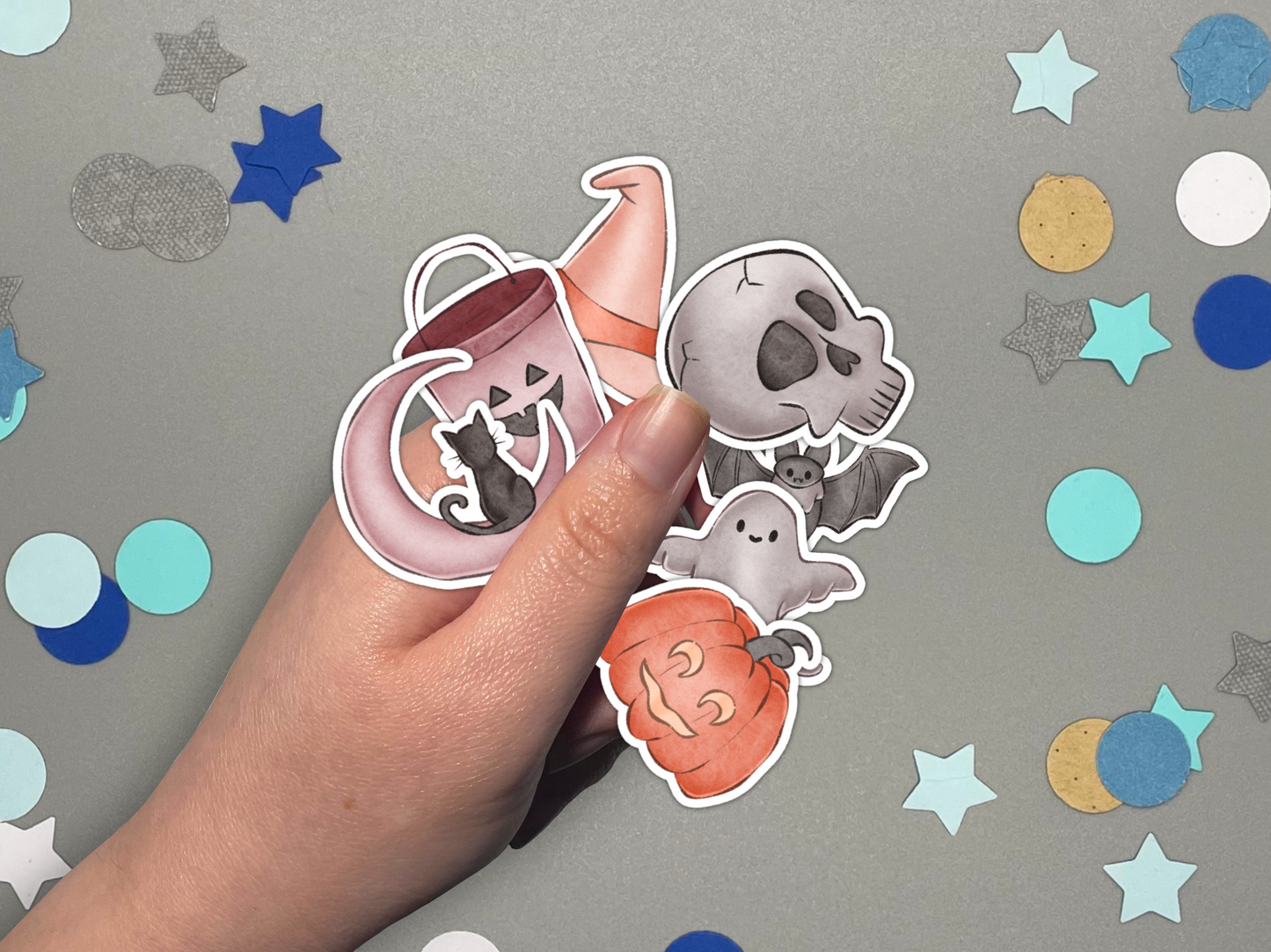 Hand holding Halloween themed stickers against a gray background with colorful star confetti