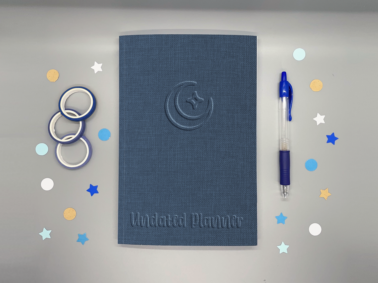 Undated planner with blue embossed-style moon and sparkle icon on a gray background surrounded by washi tape, confetti, and a blue pen.