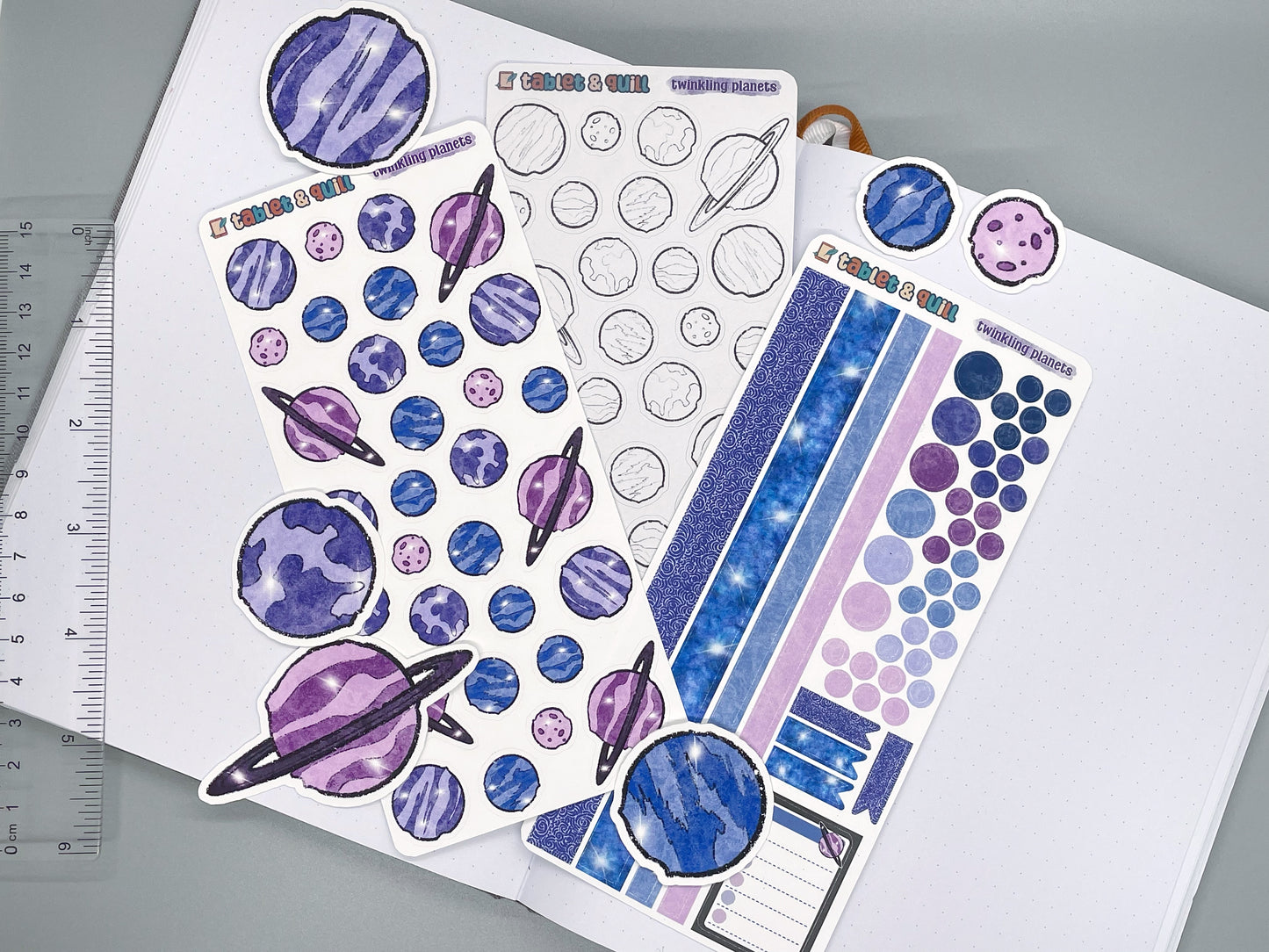 Twinkling planets sticker sheets and die cut stickers on journal with ruler on gray background.