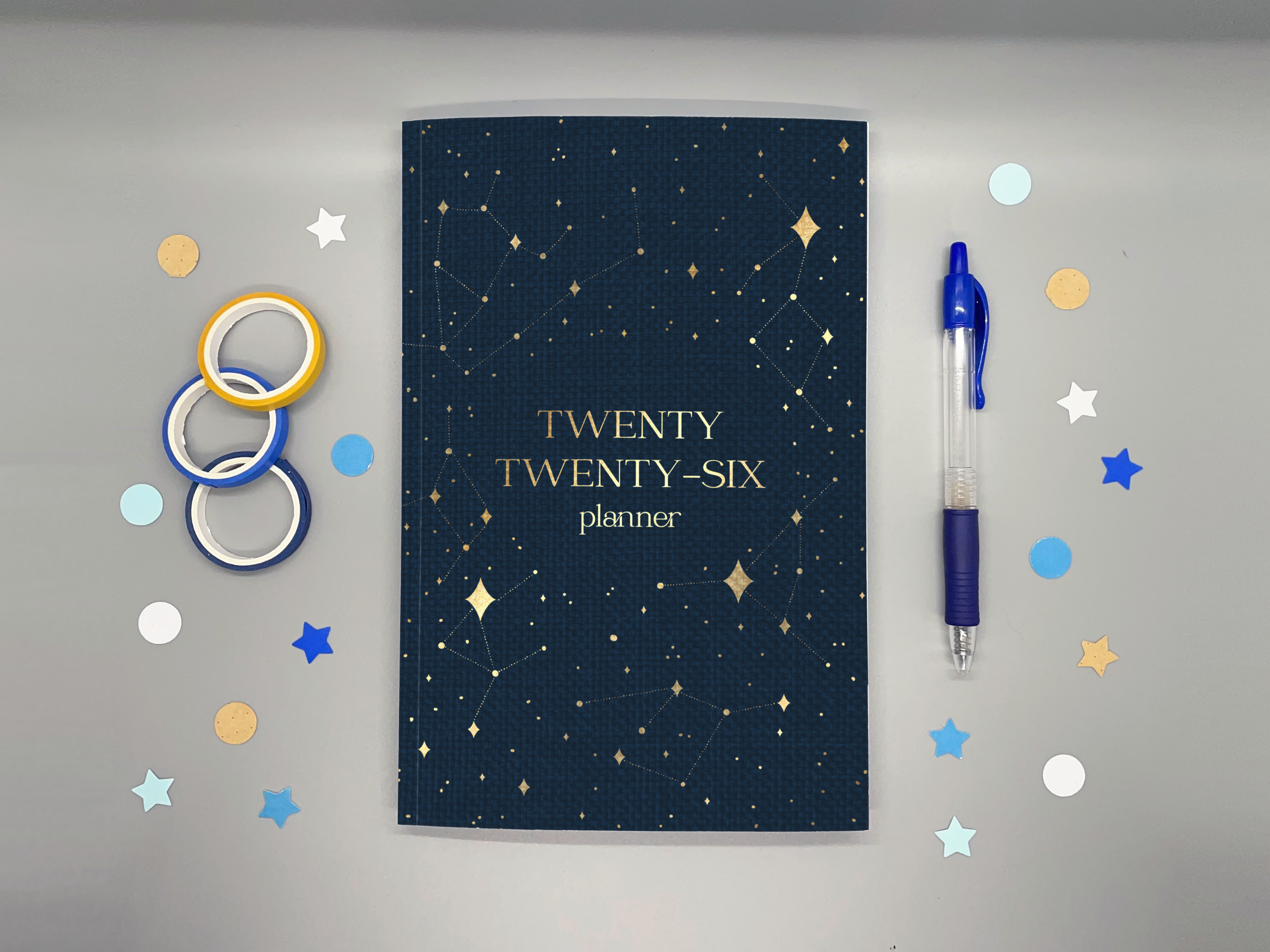 2026 planner with washi tape, confetti, and a pen around it all on a gray background.