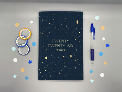 2026 planner with washi tape, confetti, and a pen around it all on a gray background.