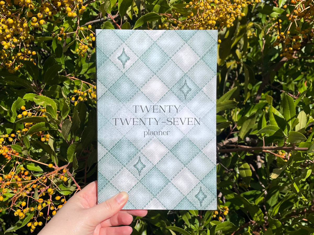 A hand holds a planner titled 'TWENTY TWENTY-SEVEN planner' with a light blue and white diamond pattern, set against a background of green leaves and yellow berries.