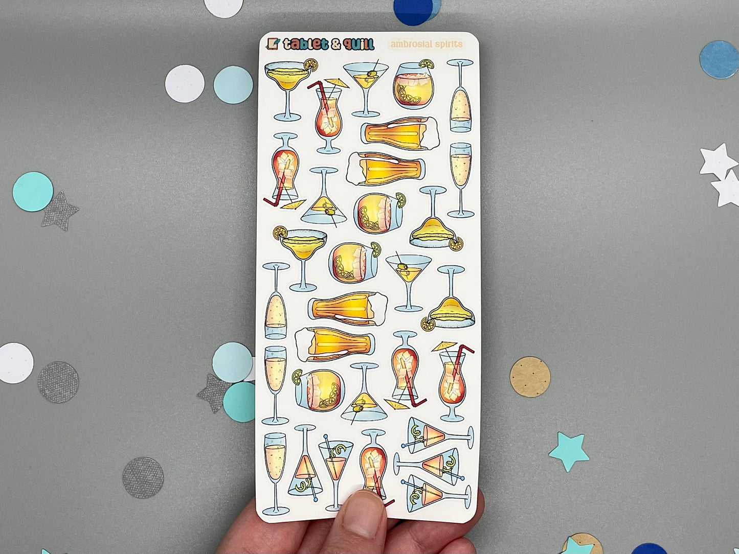 Sheet of cocktail-themed stickers held by a hand on a gray background with decorative confetti.