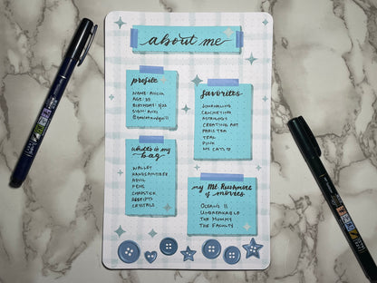 About me page decorated with blue pieces of paper on a light blue grid background. At the bottom are blue button stickers. Page is on a marbled surface with two pens.