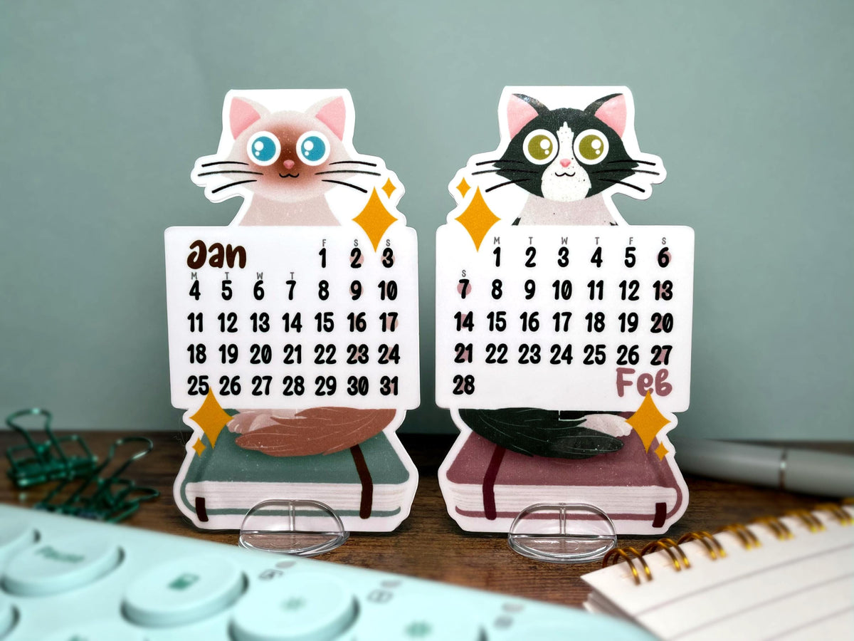 Two cute paper desk calendar cutouts shaped like cats sitting on journals, displayed on clear acrylic stands on a desk with office items in the foreground.