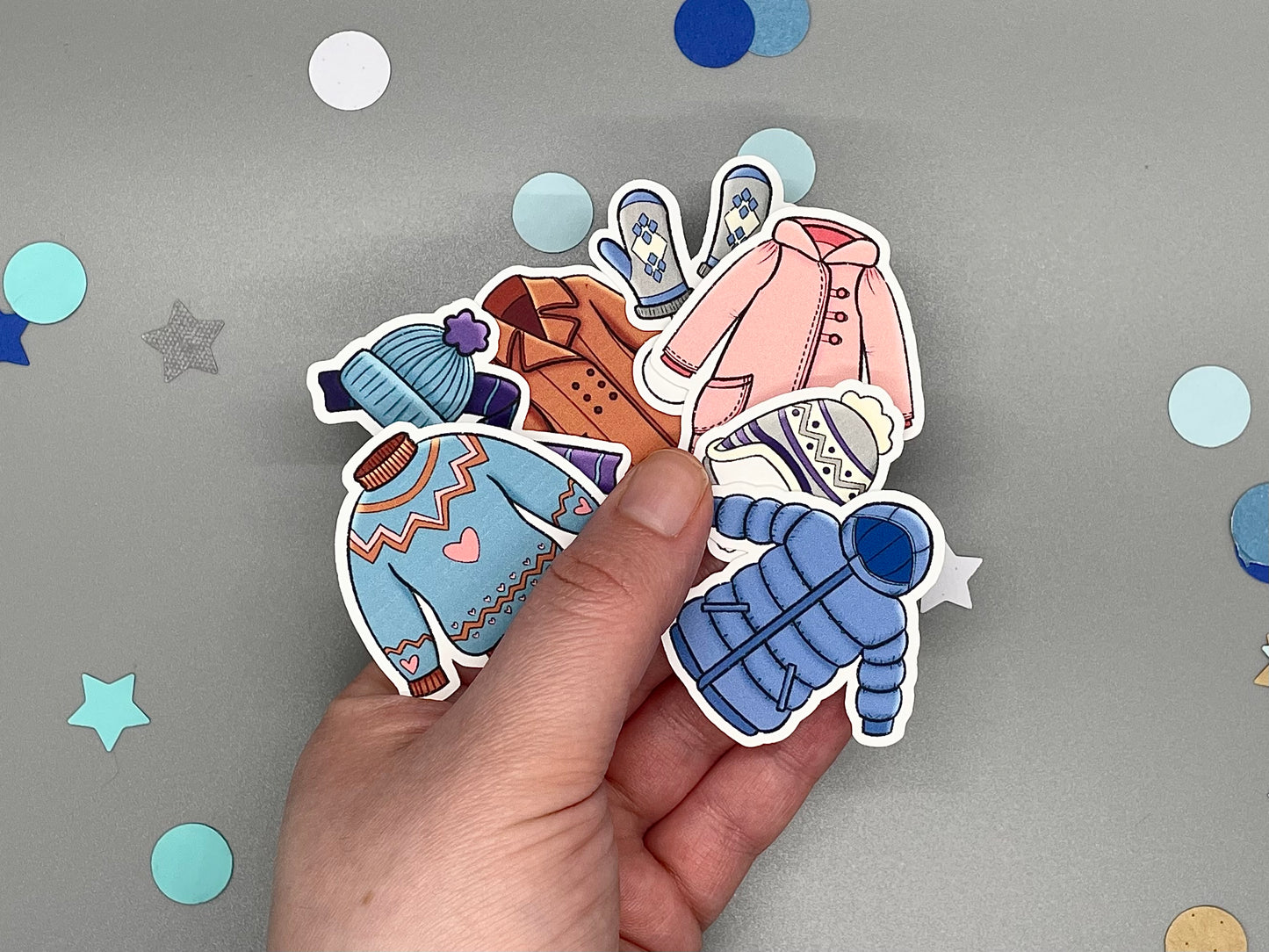 Hand holding cartoon-style clothing stickers on a gray background with colorful confetti.