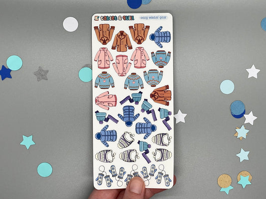 Sticker sheet of winter clothes themed stickers held by a hand on a gray background with decorative stars.