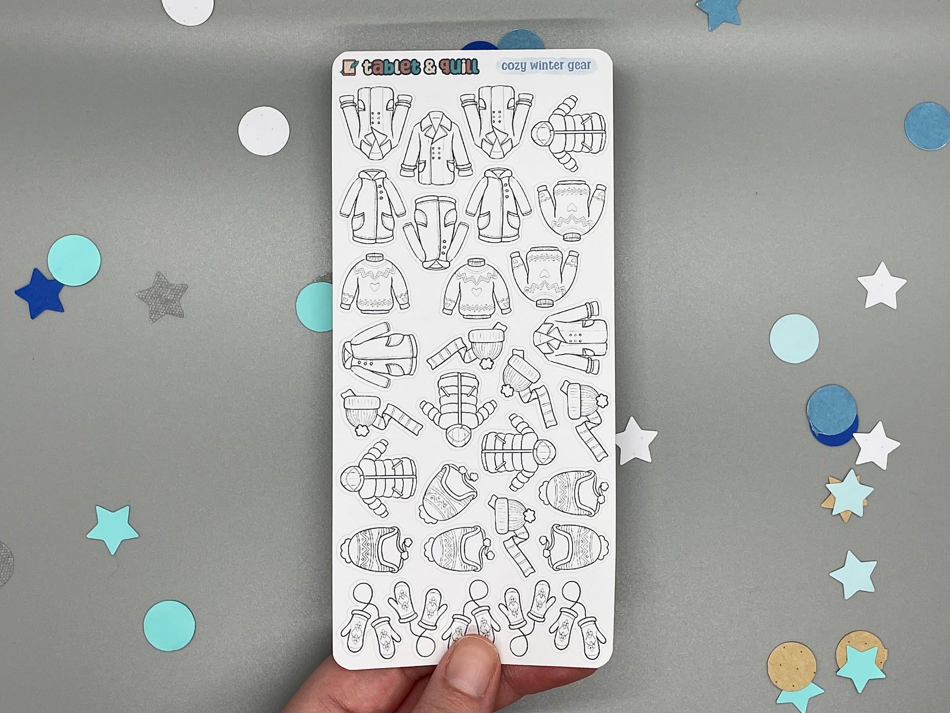 Sticker sheet of diy color stickers winter clothes themed held over a gray background with star and circle confetti.