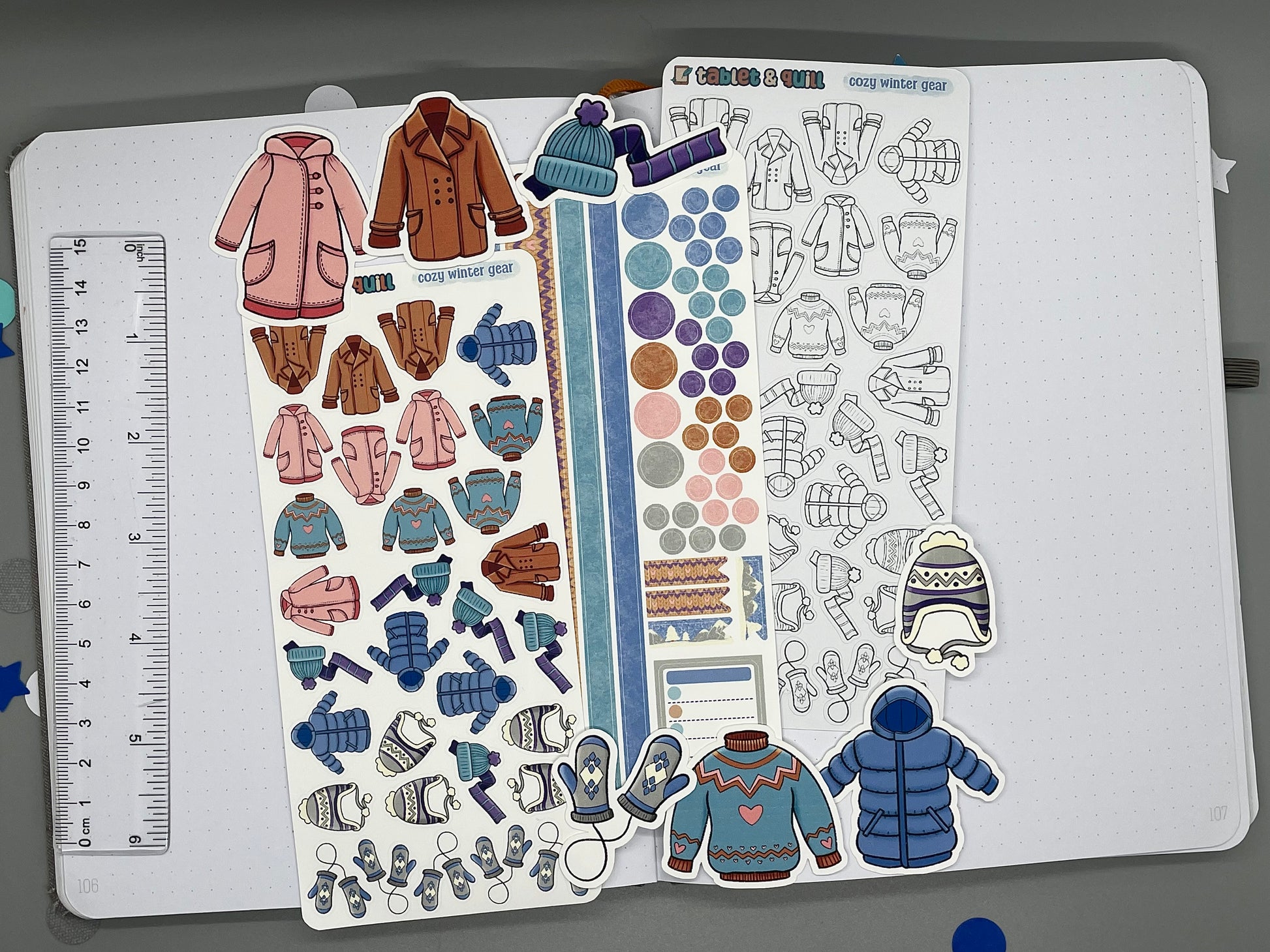Sticker sheets and die cut stickers with winter clothing designs on a notebook page with a ruler for scale.