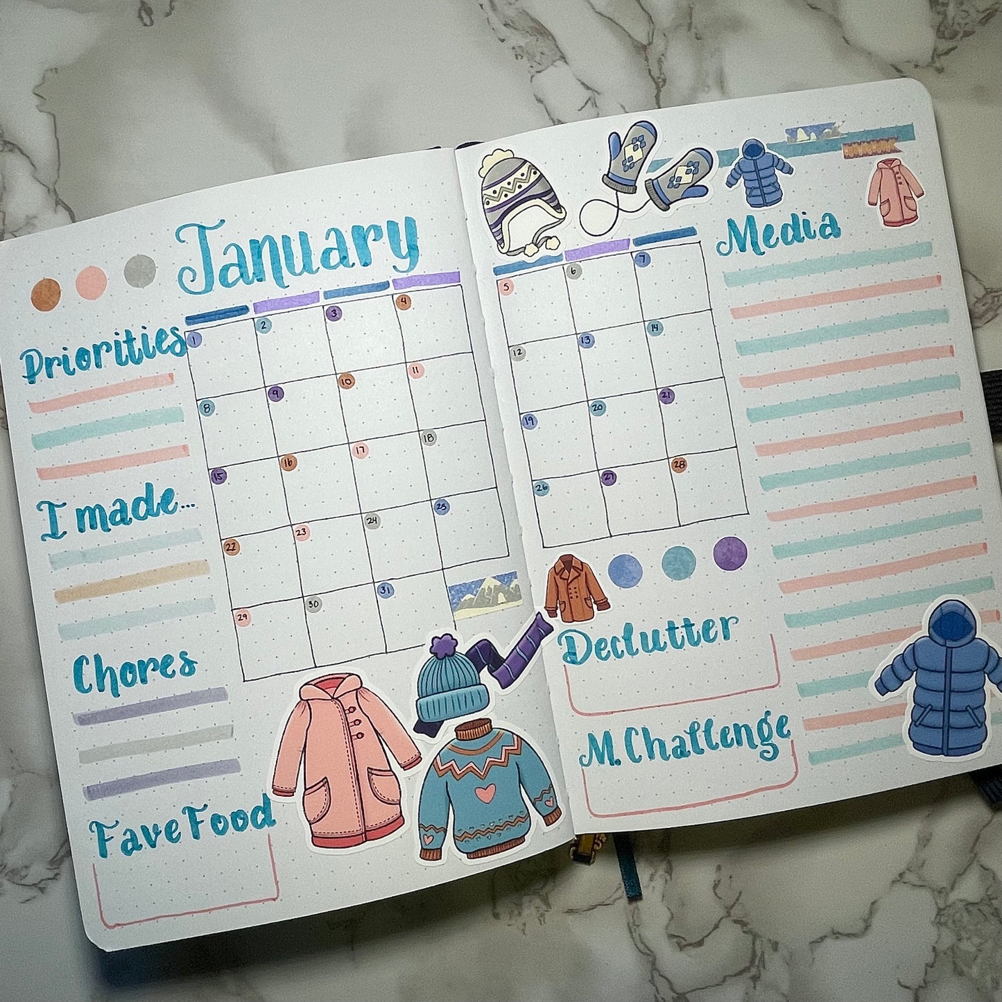Open bullet journal with January calendar and winter clothes themed stickers on a marble surface