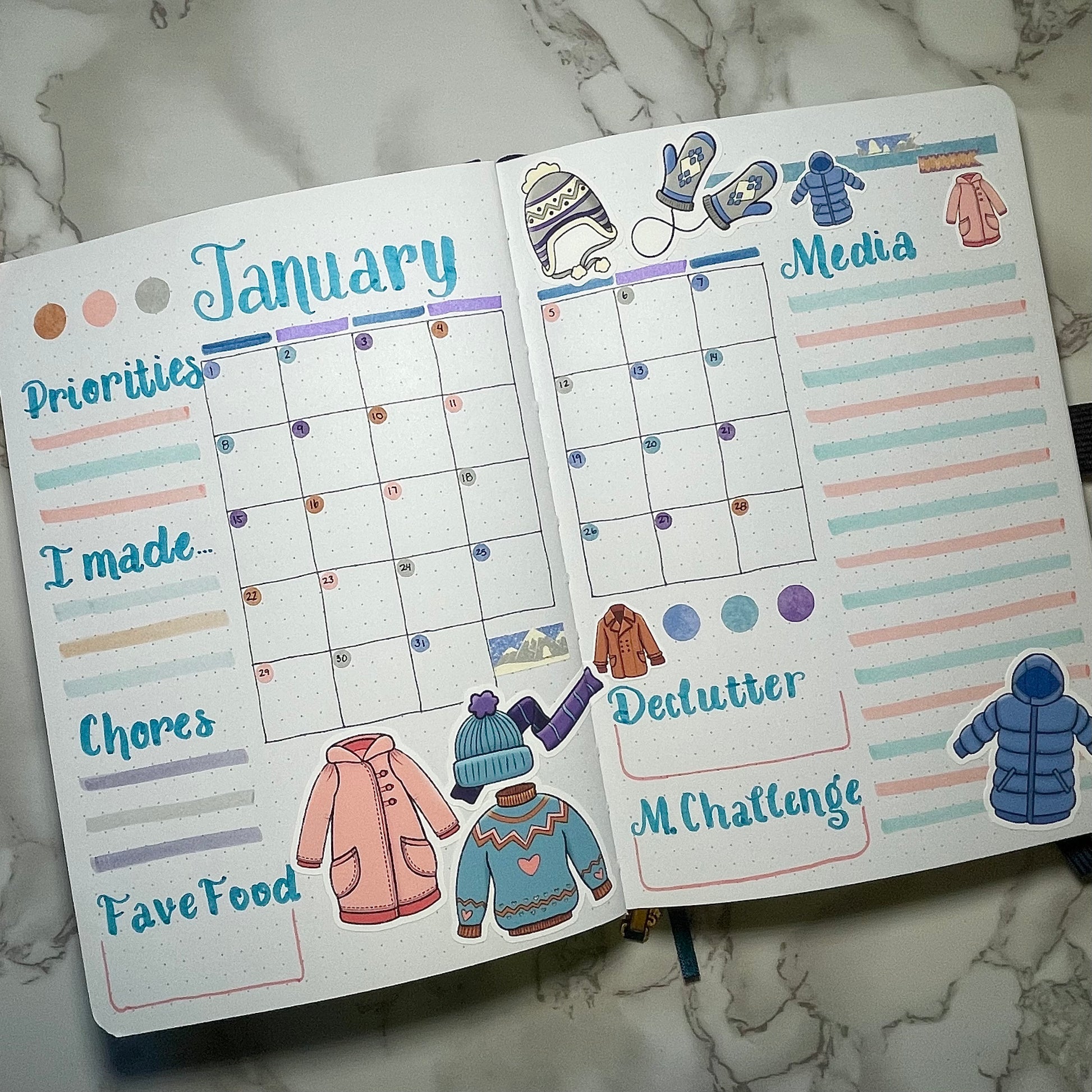 Open bullet journal with January calendar and winter clothes themed stickers on a marble surface