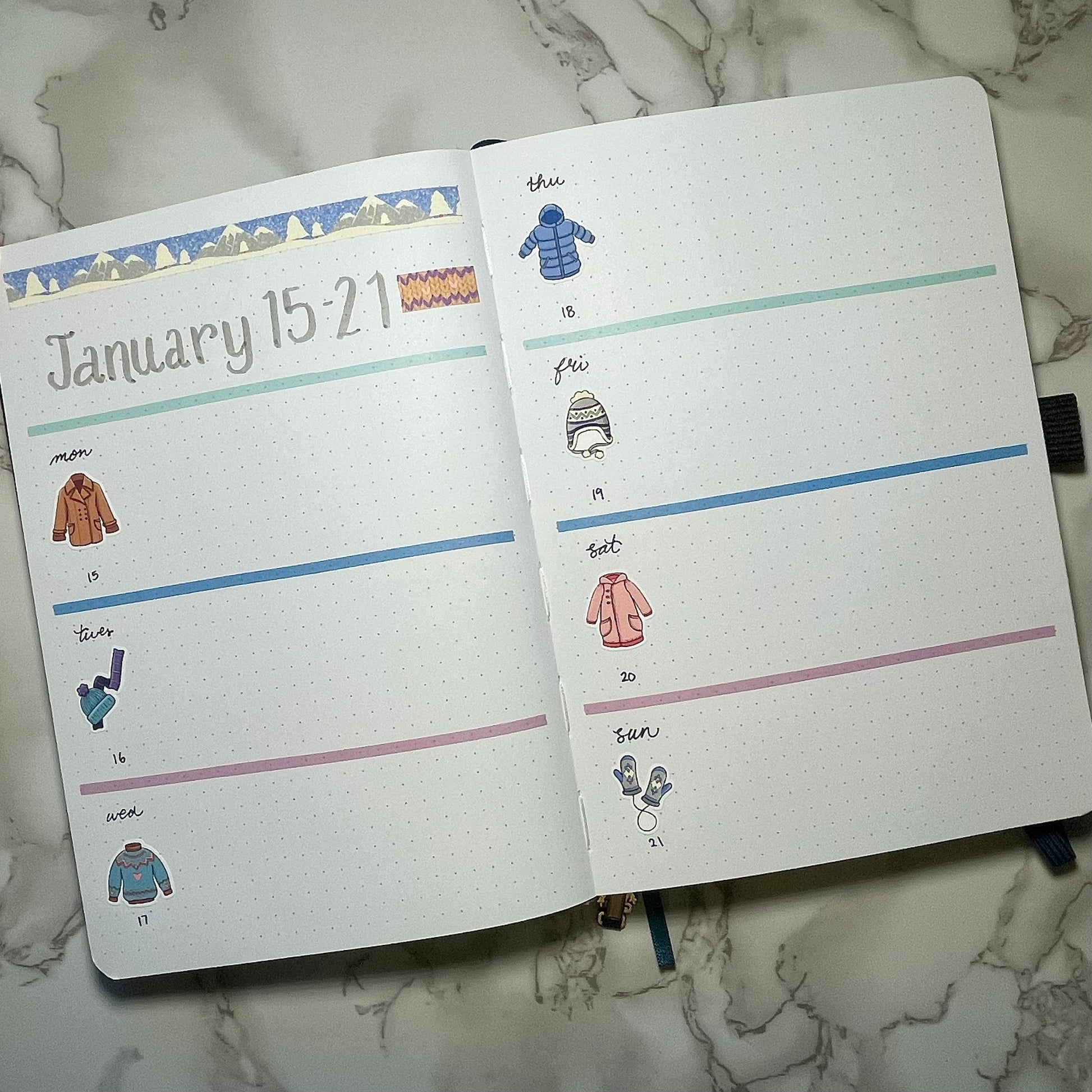 Open bullet journal with weekly spread and winter clothes themed on a marble surface.