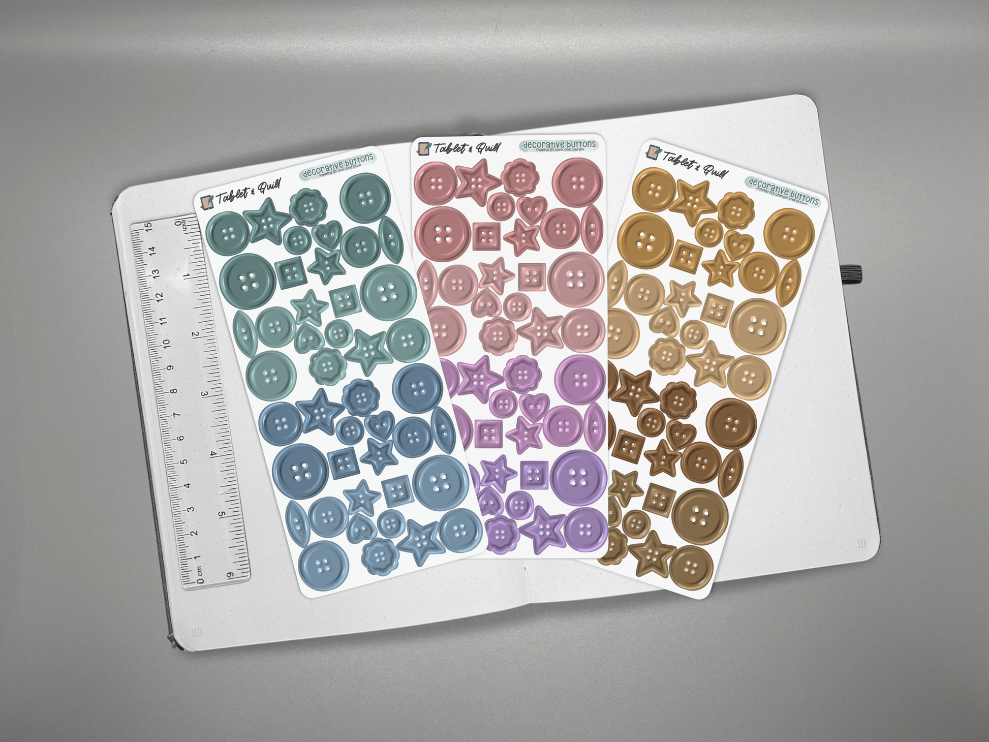 Three decorative buttons sticker sheets in different colors on an open journal with a ruler for scale on a gray background.