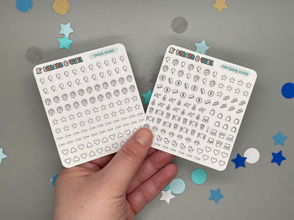 Two sticker sheets with small icons held by a hand against a gray background with decorative stars.