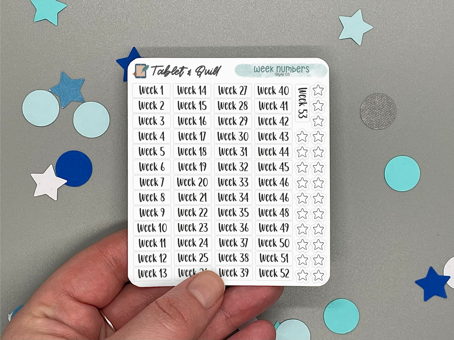 Hand holding a planner sticker sheet with week numbers on a gray background with decorative stickers.