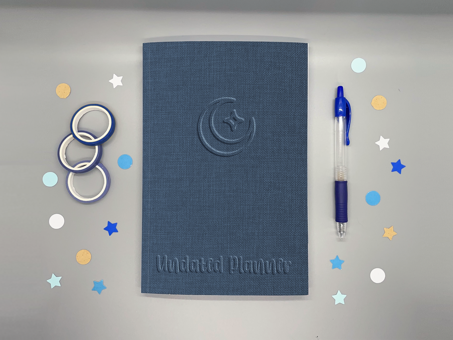 Undated planner with blue embossed-style moon and sparkle icon on a gray background surrounded by washi tape, confetti, and a blue pen.