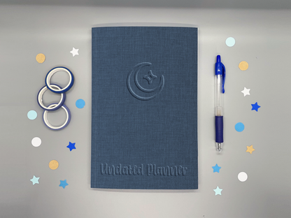 Undated planner with blue embossed-style moon and sparkle icon on a gray background surrounded by washi tape, confetti, and a blue pen.
