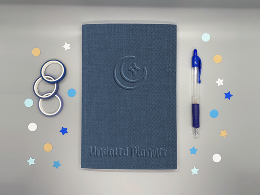 Undated planner with blue embossed-style moon and sparkle icon on a gray background surrounded by washi tape, confetti, and a blue pen.