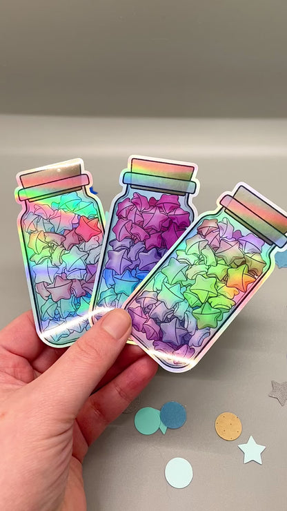 Holographic laminated pride stickers. Paper stars in a jar. A hand moves them so you can see it's holographic. Rainbow, bisexual, transgender flag colors.