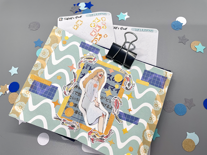 Two stars and sparkles DIY color sticker sheets on a gray background. One sheet has colored sparkles that are removed from the sheet. A small journal sits on top decorated with paper and stickers, including koi stickers colored with colored pencils and stars and sparkles colored with markers.