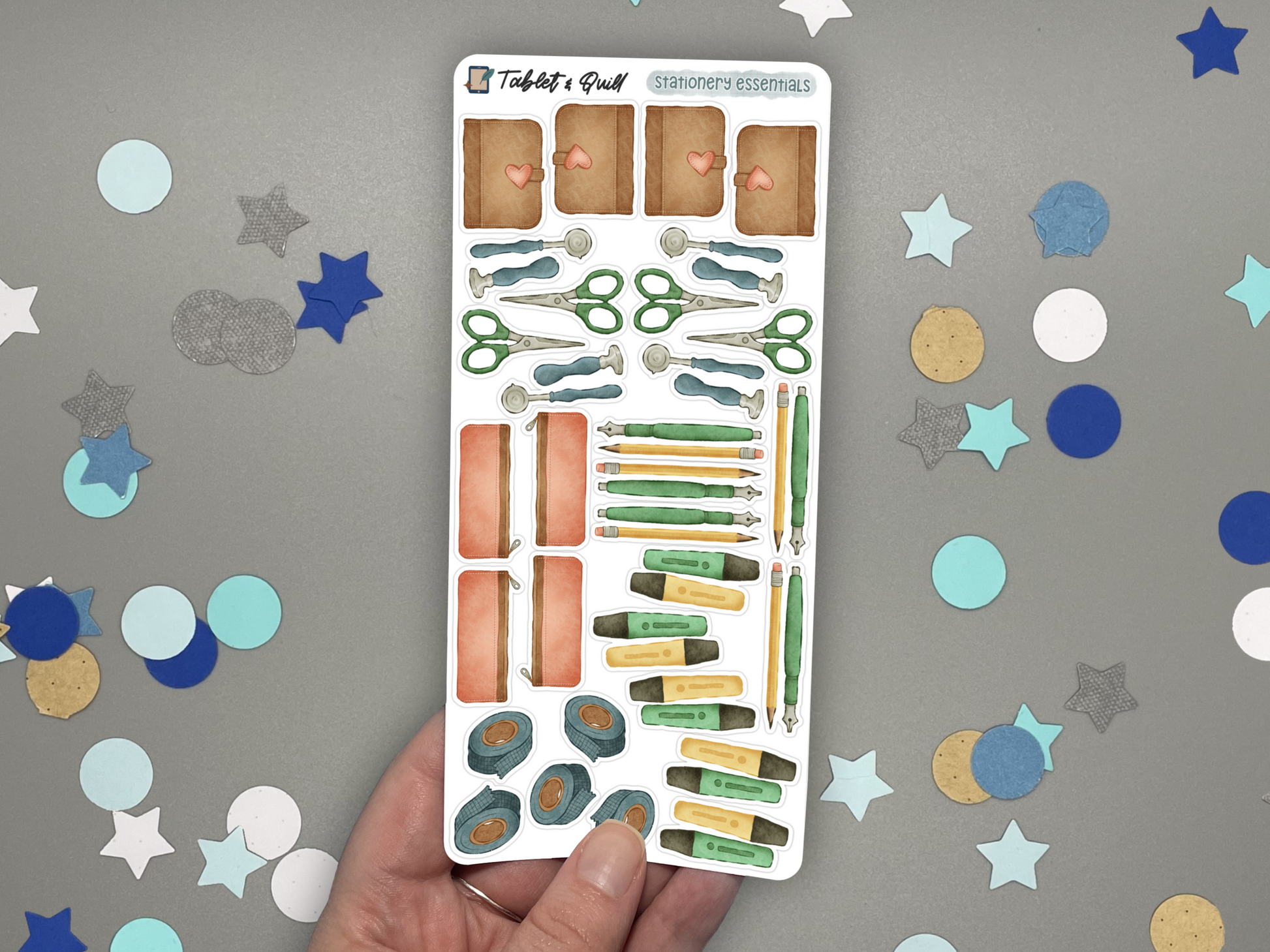 Stationery themed bullet journal stickers in color held by a hand and over a gray background with confetti. 