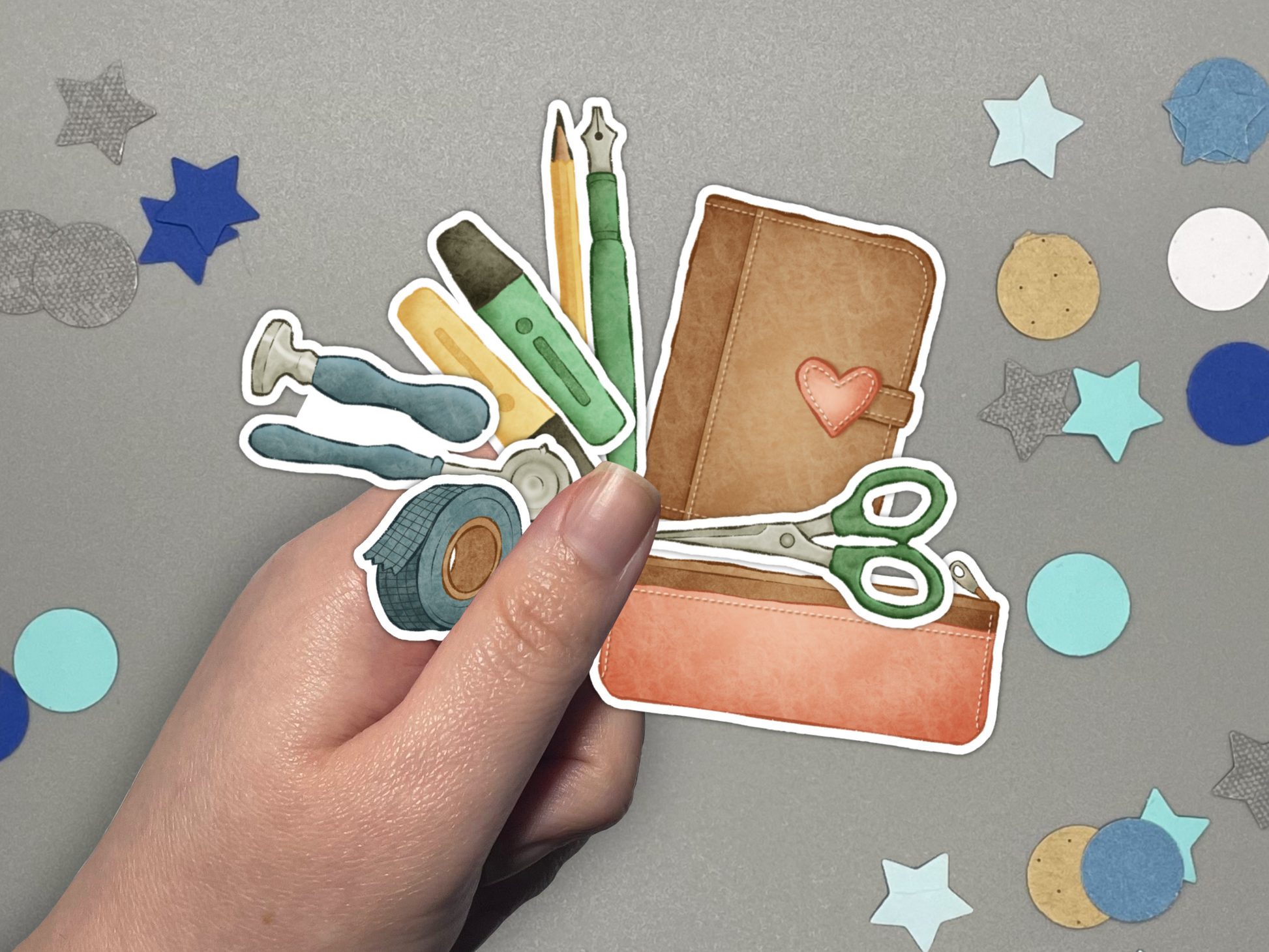Hand holding a collection of stationery themed stickers on a gray background with confetti.